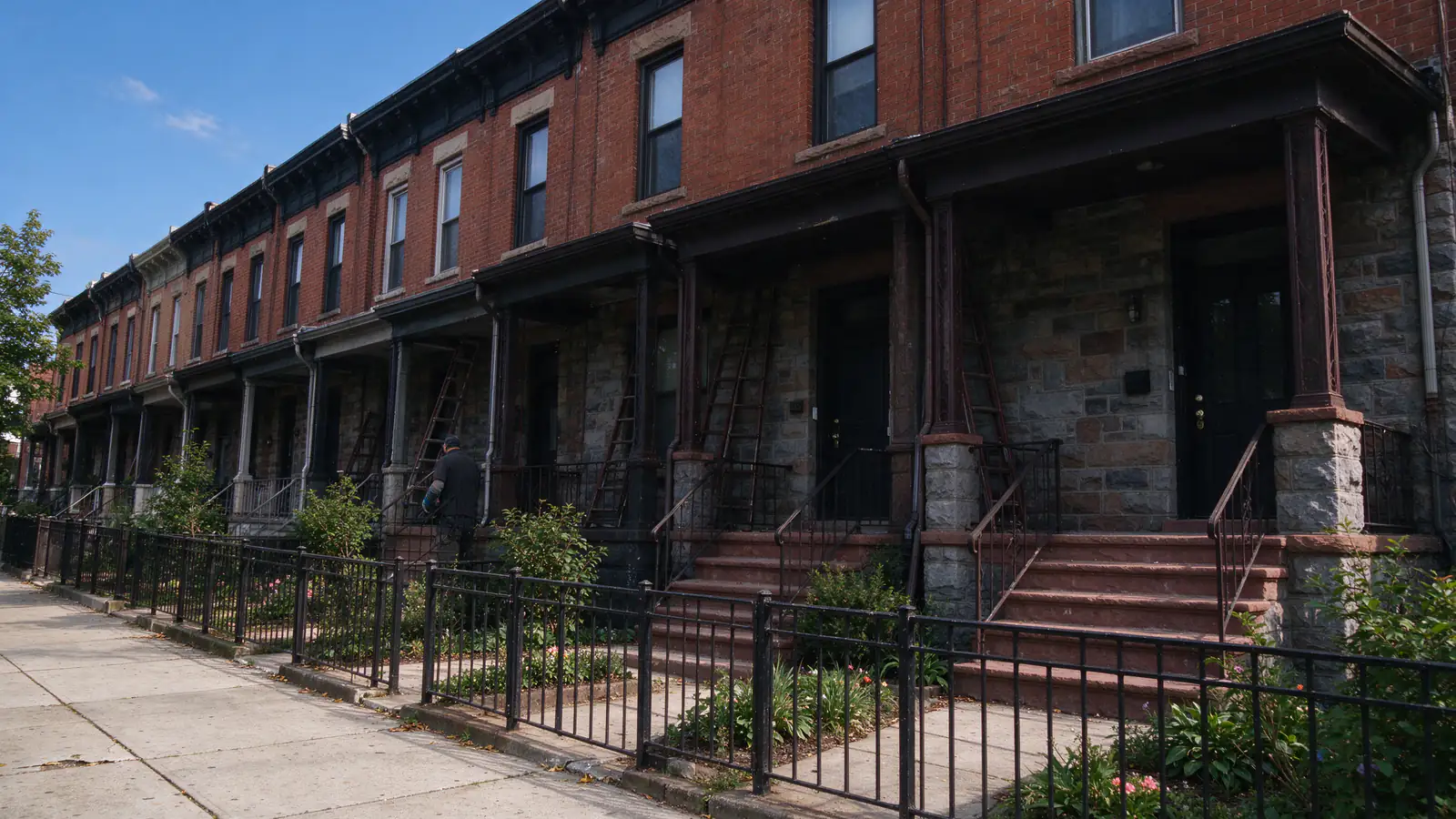 A row of brick twin houses with shared front porches on a tree-lined West Philadelphia street